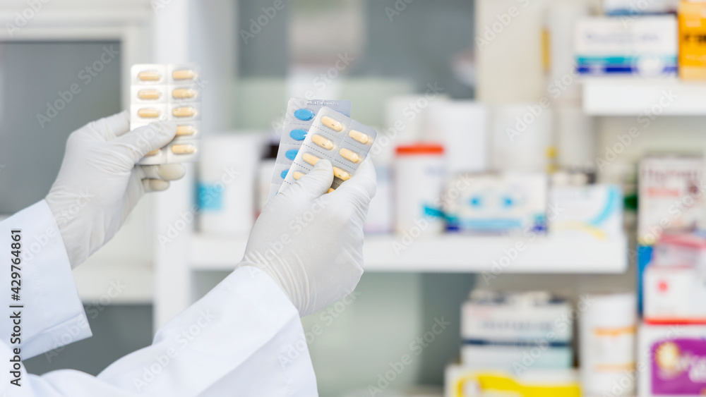 Fototapeta premium Pharmacist holding medicine box and capsule pack in pharmacy drugstore.Pharmacist holding capsule pack with shelves background.Hand holding medicine capsule pack at the pharmacy drugstore.