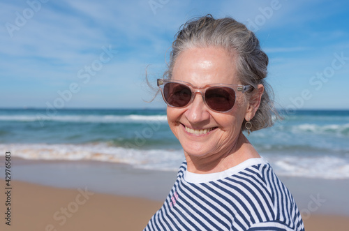 Head and shoulders portrait of senior woman smiling with sunglasses at beach (selective focus)