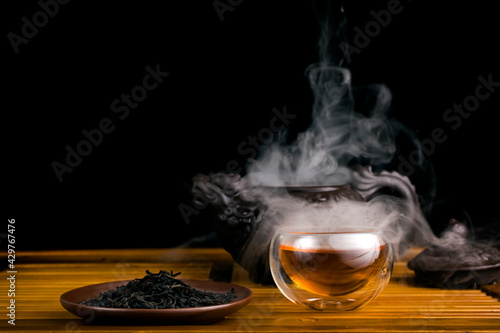 Chinese tea ceremony. Clay teapot with red tea Lapsang souchong on a black background, heap of tea and glass thermo cup of hot tea with vapour. photo