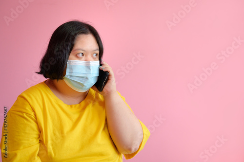 Image of a handicapped woman with Down Syndrome learning with a mobile phone making gestures on the phone She is a disabled student with Down syndrome wearing a medical mask on the pink background.