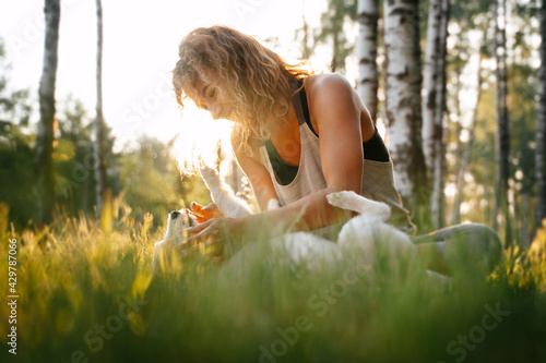 Beautiful young woman with curly hair playing with her little friend dog jack russell in the park outdoors at sunrise. Lifestyle portrait with pet on a walk.