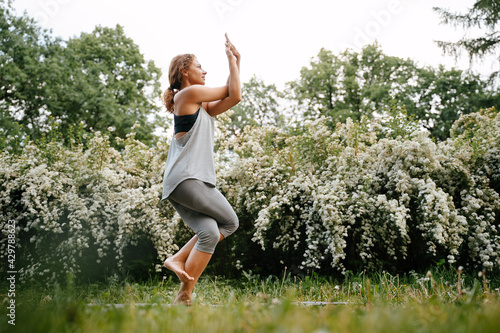 Athletic woman practices yoga and stands on one leg on a yoga mat in the park. Eka Pada Utkatasana pose.