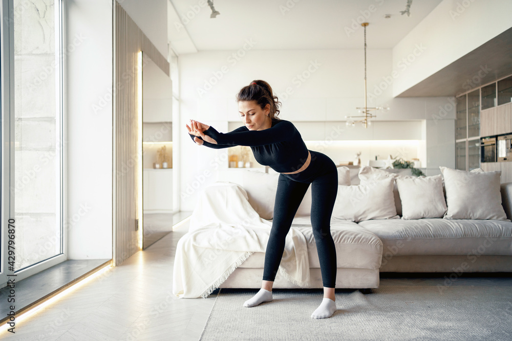 A brunette woman does a workout in a bright apartment. Body stretching exercises for the cardio system. Healthy lifestyle. Stylish sports black suit is tight-fitting. A brunette woman does a workout in a bright apartment. Body stretching exercises for the cardio system. Healthy lifestyle. Stylish sports black suit is tight-fitting.