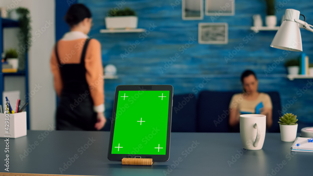 Home studio equipped with tablet computer with mock up green screen chroma key display standing in vertical mode on office desk. In background two collegues talking about internet advertising Home studio equipped with tablet computer with mock up green screen chroma key display standing in vertical mode on office desk. In background two collegues talking about internet advertising