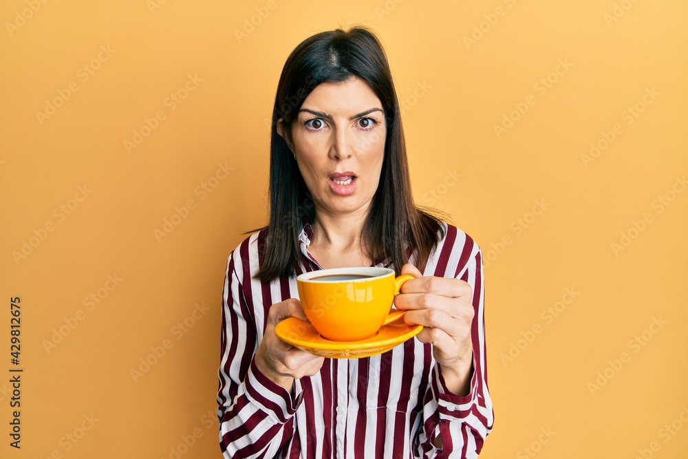 Young hispanic woman drinking a cup of coffee in shock face, looking skeptical and sarcastic, surprised with open mouth Young hispanic woman drinking a cup of coffee in shock face, looking skeptical and sarcastic, surprised with open mouth