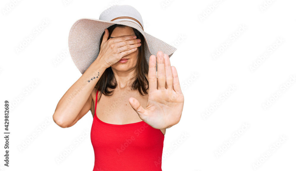 Beautiful brunette woman wearing swimsuit and summer hat covering eyes with hands and doing stop gesture with sad and fear expression. embarrassed and negative concept. Beautiful brunette woman wearing swimsuit and summer hat covering eyes with hands and doing stop gesture with sad and fear expression. embarrassed and negative concept.