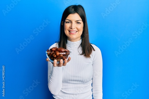 Young hispanic woman holding dates bowl looking positive and happy standing and smiling with a confident smile showing teeth