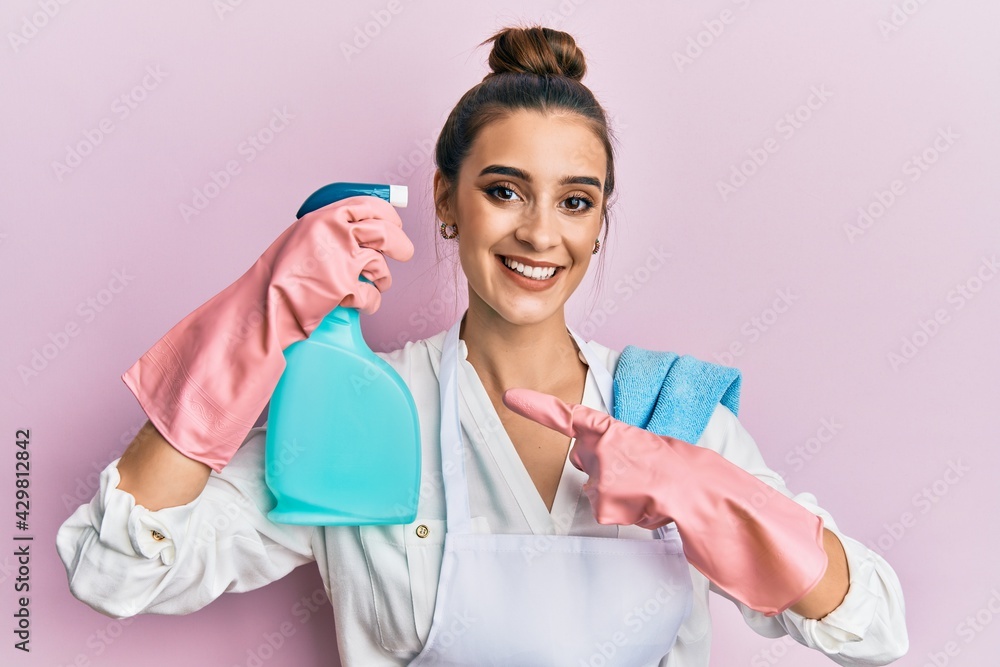 Beautiful brunette young woman wearing apron holding cleaning spray pointing to head smiling happy pointing with hand and finger Beautiful brunette young woman wearing apron holding cleaning spray pointing to head smiling happy pointing with hand and finger