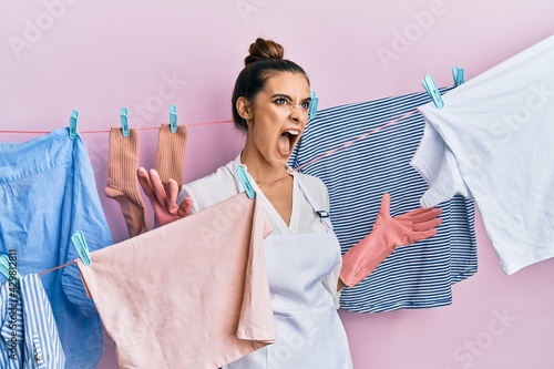 Beautiful brunette young woman washing clothes at clothesline crazy and mad shouting and yelling with aggressive expression and arms raised. frustration concept.