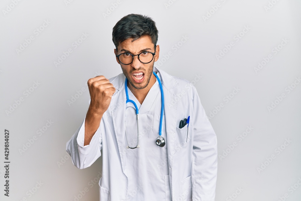 Young handsome man wearing doctor uniform and stethoscope angry and mad raising fist frustrated and furious while shouting with anger. rage and aggressive concept. Young handsome man wearing doctor uniform and stethoscope angry and mad raising fist frustrated and furious while shouting with anger. rage and aggressive concept.