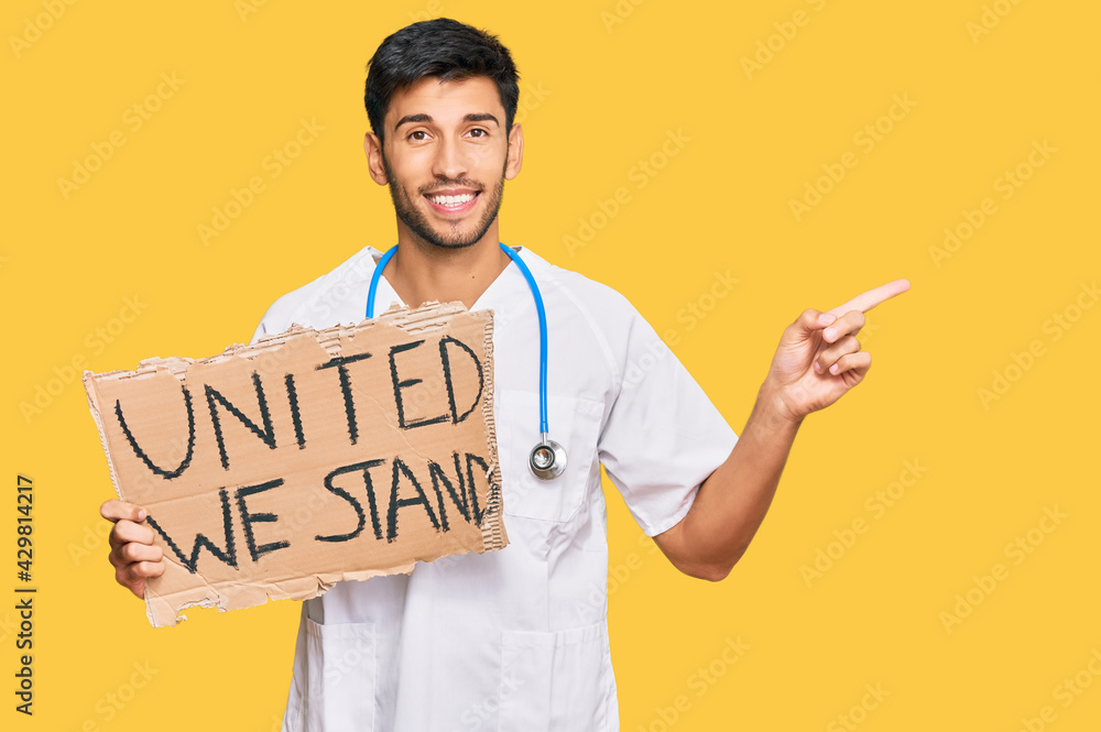Young handsome man wearing doctor uniform holding united we stand banner smiling happy pointing with hand and finger to the side Young handsome man wearing doctor uniform holding united we stand banner smiling happy pointing with hand and finger to the side