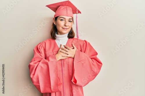 Young caucasian woman wearing graduation cap and ceremony robe smiling with hands on chest with closed eyes and grateful gesture on face. health concept.