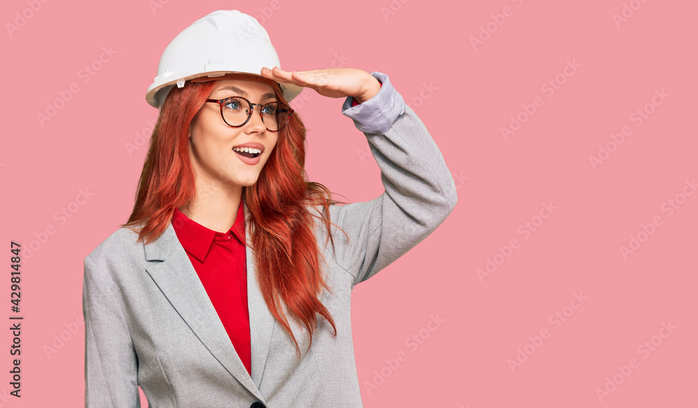 Young redhead woman wearing architect hardhat very happy and smiling looking far away with hand over head. searching concept. Young redhead woman wearing architect hardhat very happy and smiling looking far away with hand over head. searching concept.