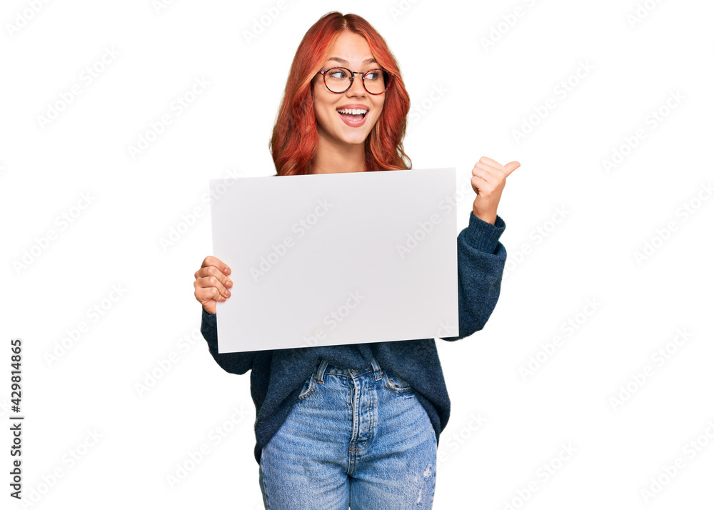 Young redhead woman holding blank empty banner pointing thumb up to the side smiling happy with open mouth Young redhead woman holding blank empty banner pointing thumb up to the side smiling happy with open mouth