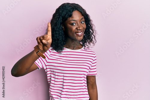 Beautiful african young woman wearing casual striped t shirt smiling with an idea or question pointing finger up with happy face, number one © Krakenimages.com