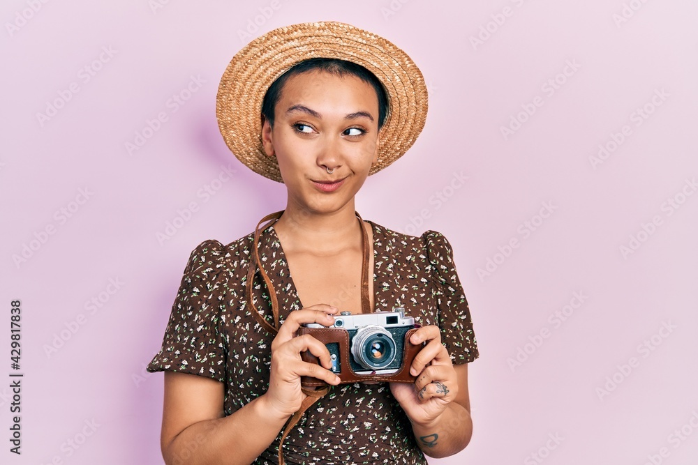 Beautiful hispanic woman with short hair wearing summer hat holding vintage camera smiling looking to the side and staring away thinking.