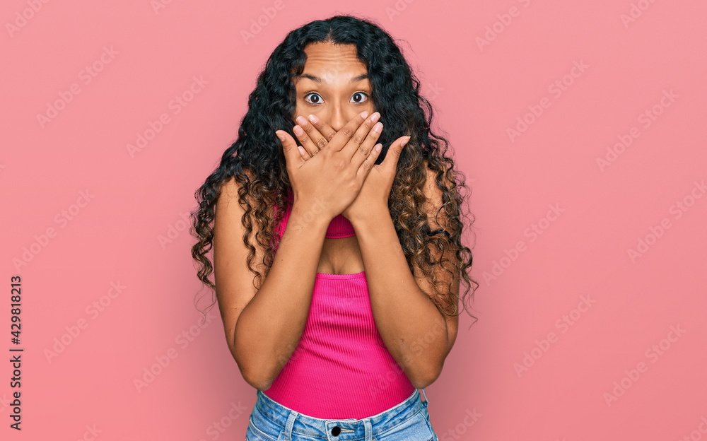 Young hispanic woman with curly hair wearing pink top shocked covering mouth with hands for mistake. secret concept. Young hispanic woman with curly hair wearing pink top shocked covering mouth with hands for mistake. secret concept.