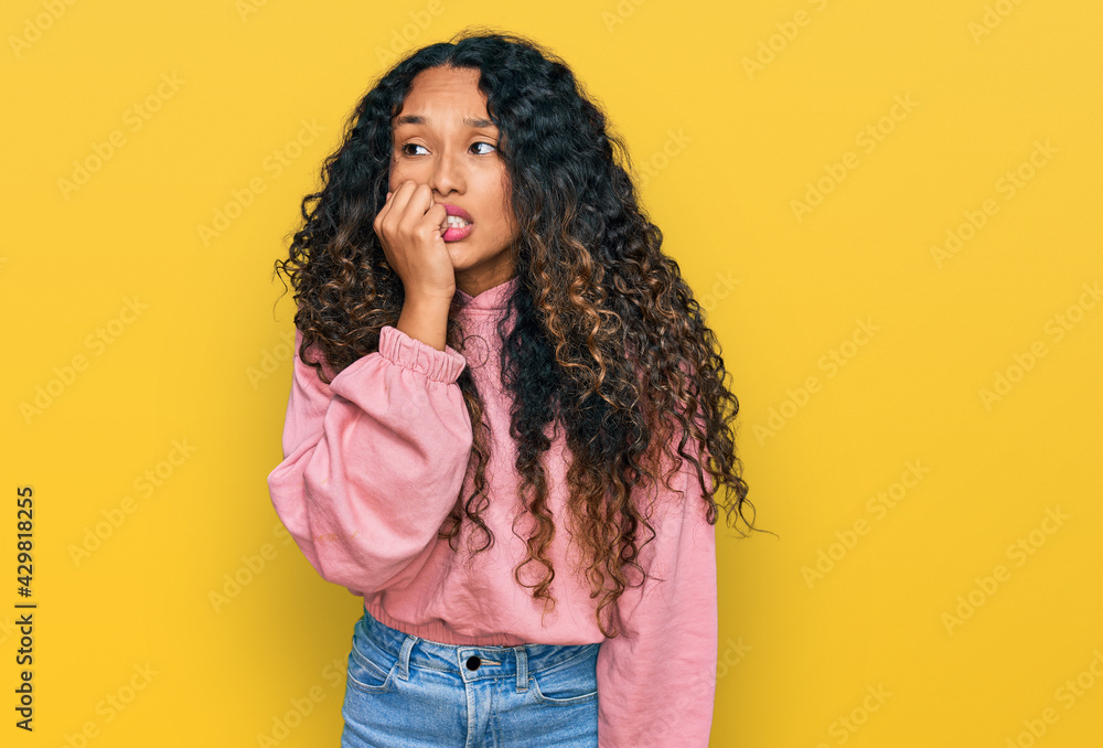 Young hispanic woman with curly hair wearing casual sweatshirt looking stressed and nervous with hands on mouth biting nails. anxiety problem. Young hispanic woman with curly hair wearing casual sweatshirt looking stressed and nervous with hands on mouth biting nails. anxiety problem.