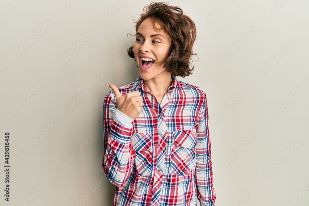 Young brunette woman wearing casual clothes pointing thumb up to the side smiling happy with open mouth Young brunette woman wearing casual clothes pointing thumb up to the side smiling happy with open mouth