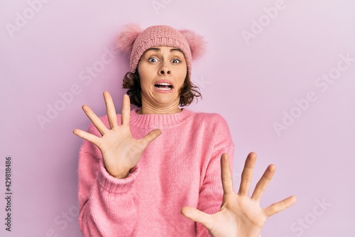 Young brunette woman wearing cute wool cap afraid and terrified with fear expression stop gesture with hands  shouting in shock. panic concept.