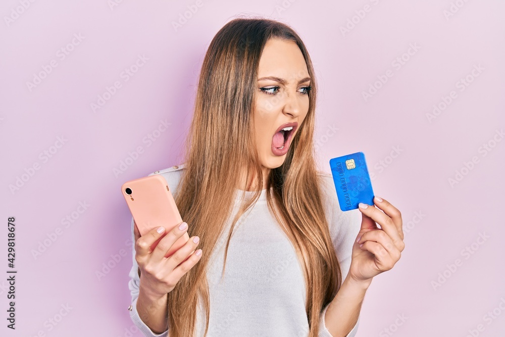 Young blonde girl holding smartphone and credit card angry and mad screaming frustrated and furious, shouting with anger. rage and aggressive concept. Young blonde girl holding smartphone and credit card angry and mad screaming frustrated and furious, shouting with anger. rage and aggressive concept.