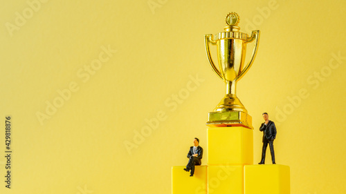Miniature of businessman stands on podium ladder with golden trophy on yellow background