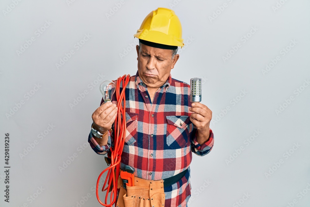 Senior hispanic man wearing handyman uniform holding led lightbulb and incandescent bulb depressed and worry for distress, crying angry and afraid. sad expression.
