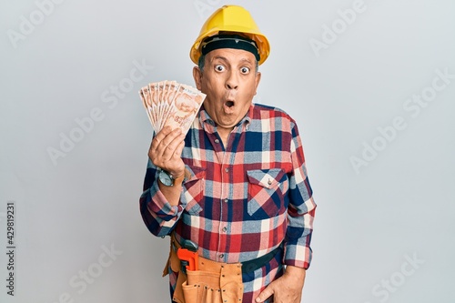 Senior hispanic man wearing handyman uniform holding turkish liras scared and amazed with open mouth for surprise, disbelief face
