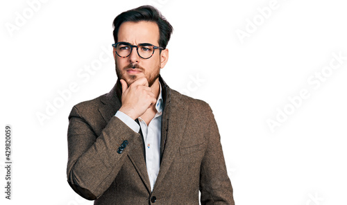 Young hispanic man wearing business jacket and glasses with hand on chin thinking about question, pensive expression. smiling with thoughtful face. doubt concept.