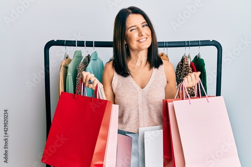 Middle age brunette personal shopper woman holding shopping bags winking looking at the camera with sexy expression, cheerful and happy face.