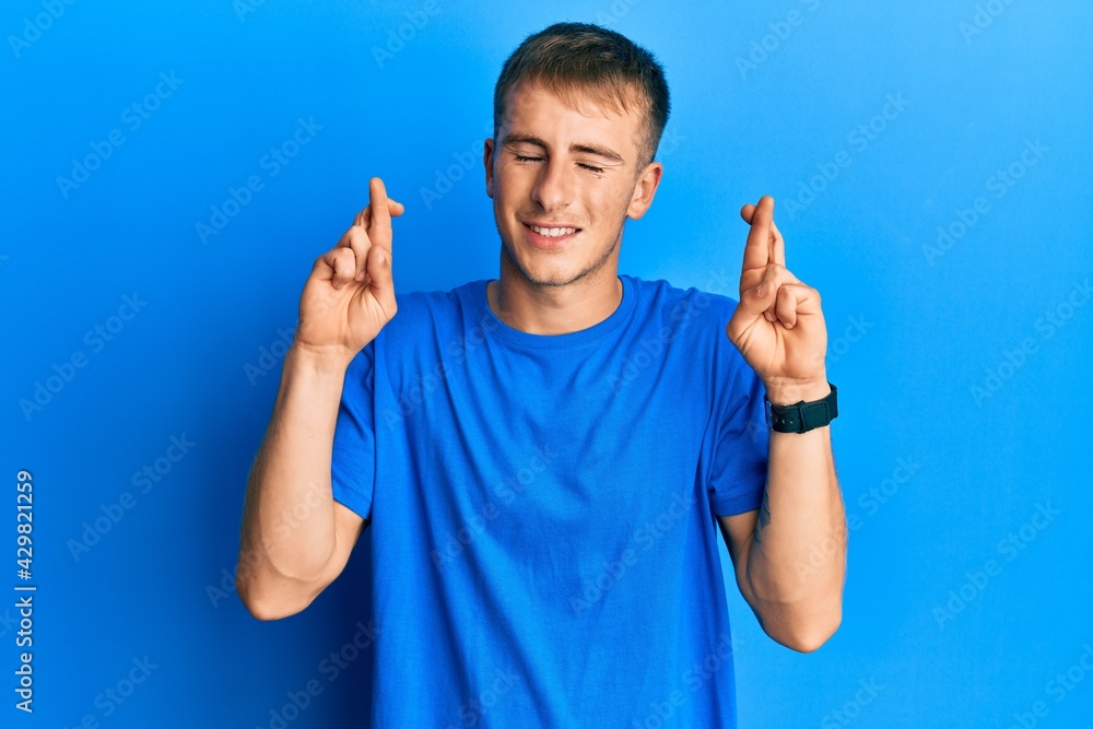 Fototapeta premium Young caucasian man wearing casual blue t shirt gesturing finger crossed smiling with hope and eyes closed. luck and superstitious concept.