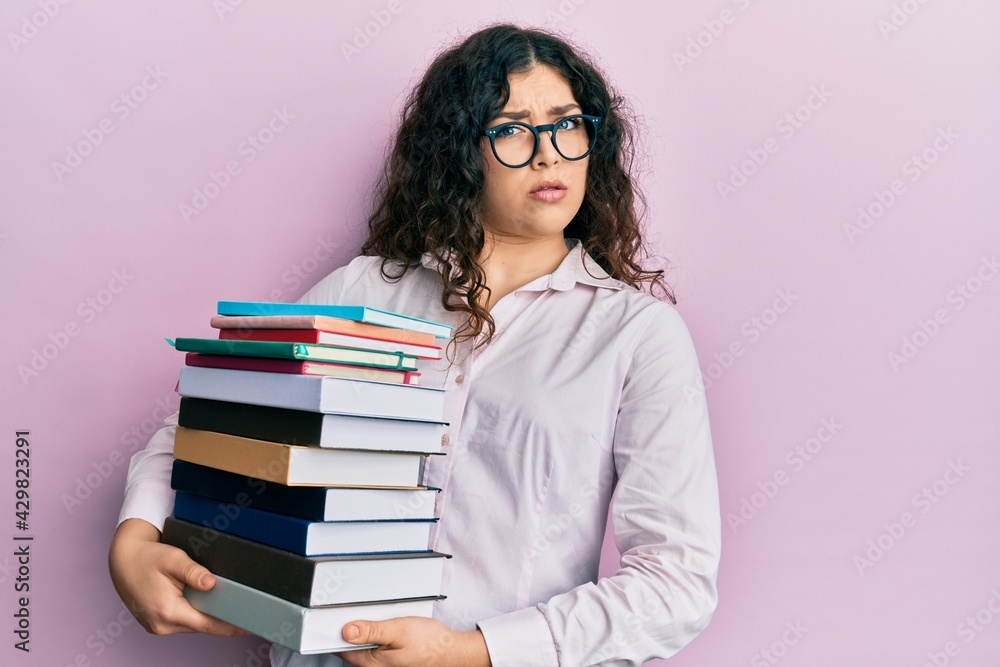 Young brunette woman with curly hair holding a pile of books in shock face, looking skeptical and sarcastic, surprised with open mouth Young brunette woman with curly hair holding a pile of books in shock face, looking skeptical and sarcastic, surprised with open mouth
