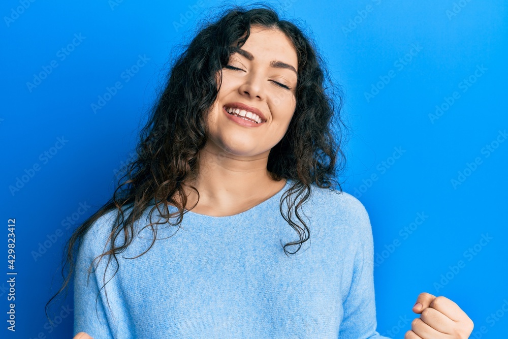Fototapeta premium Young brunette woman with curly hair wearing casual clothes very happy and excited doing winner gesture with arms raised, smiling and screaming for success. celebration concept.