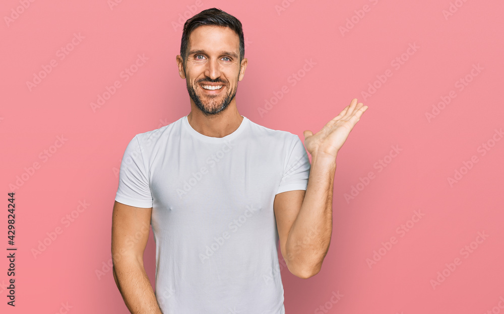 Handsome man with beard wearing casual white t shirt smiling cheerful presenting and pointing with palm of hand looking at the camera. Handsome man with beard wearing casual white t shirt smiling cheerful presenting and pointing with palm of hand looking at the camera.