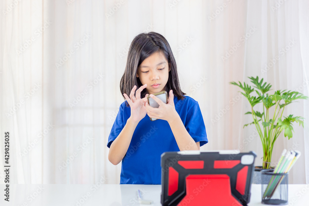 Asian kid standing and using a smartphone, Girls study online on a tablet while in the living room, kids and education concepts Asian kid standing and using a smartphone, Girls study online on a tablet while in the living room, kids and education concepts