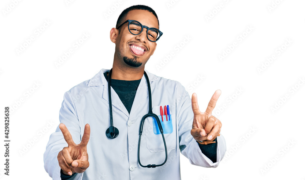 Young african american man wearing doctor uniform and stethoscope smiling with tongue out showing fingers of both hands doing victory sign. number two.