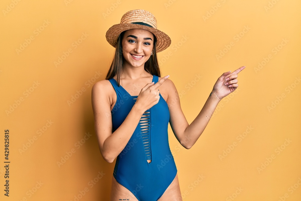 Young brunette girl wearing swimsuit and summer hat smiling and looking at the camera pointing with two hands and fingers to the side. Young brunette girl wearing swimsuit and summer hat smiling and looking at the camera pointing with two hands and fingers to the side.