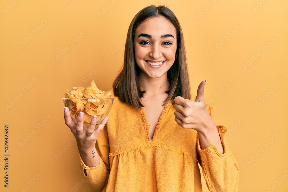 Young brunette girl holding bowl with spaghetti smiling happy and positive, thumb up doing excellent and approval sign Young brunette girl holding bowl with spaghetti smiling happy and positive, thumb up doing excellent and approval sign