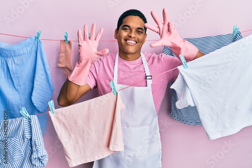 Young handsome hispanic man wearing cleaner apron holding clothes on clothesline showing and pointing up with fingers number nine while smiling confident and happy.