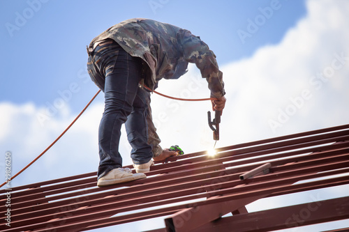 Wallpaper Mural Red painted steel roof truss, with roof welders working at dangerous heights. Welding of steel is very technically demanding and is only professional. Bright sky background Torontodigital.ca