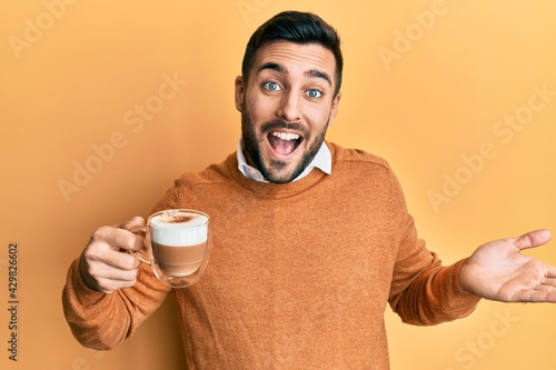 Young hispanic man holding coffee celebrating achievement with happy smile and winner expression with raised hand