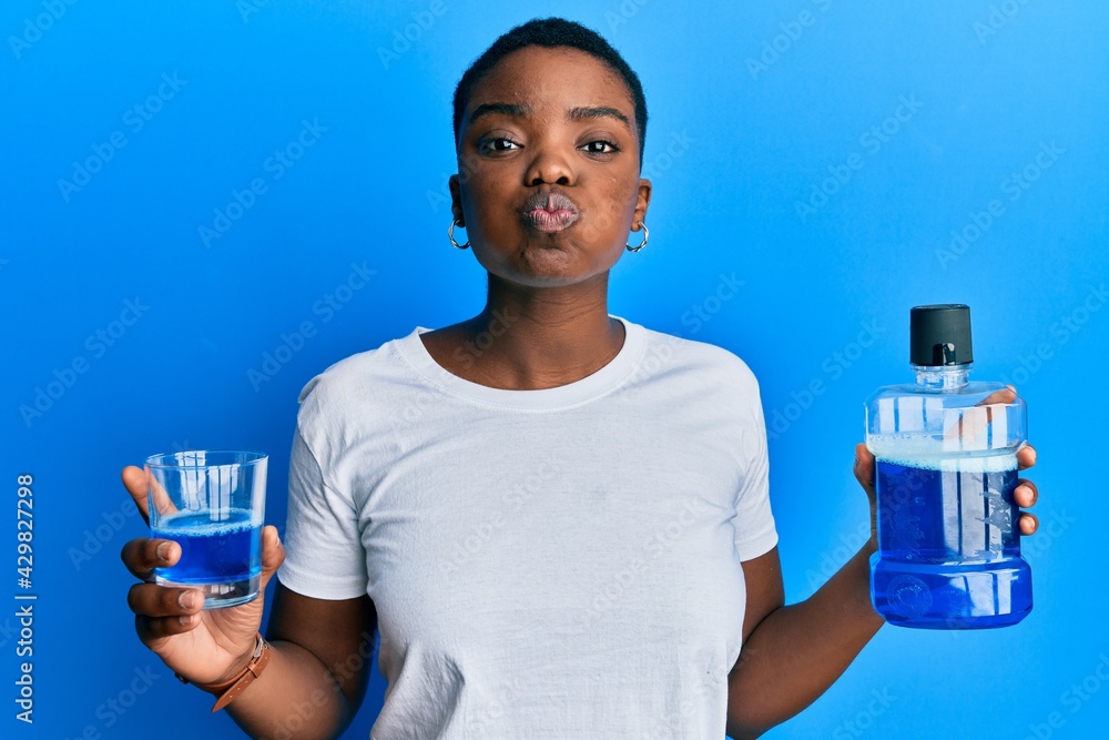 Young african american woman holding mouthwash for fresh breath puffing cheeks with funny face. mouth inflated with air, catching air. Young african american woman holding mouthwash for fresh breath puffing cheeks with funny face. mouth inflated with air, catching air.