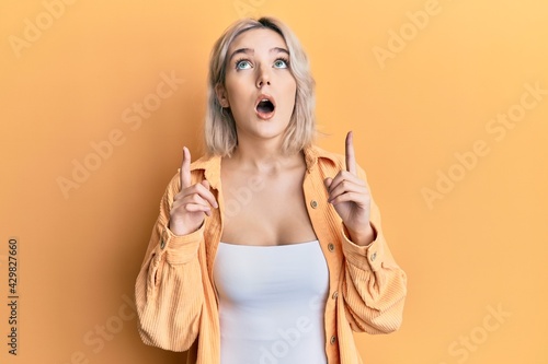 Young blonde girl wearing casual clothes amazed and surprised looking up and pointing with fingers and raised arms.