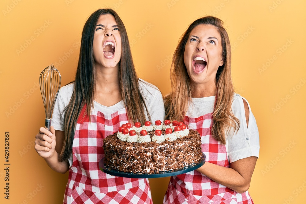 Hispanic family of mother and daughter wearing baker apron holding homemade cake angry and mad screaming frustrated and furious, shouting with anger looking up.