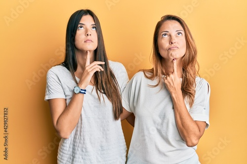 Hispanic family of mother and daughter wearing casual white tshirt thinking concentrated about doubt with finger on chin and looking up wondering