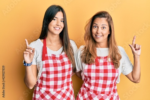 Hispanic family of mother and daughter wearing baker uniform over yellow background surprised with an idea or question pointing finger with happy face, number one