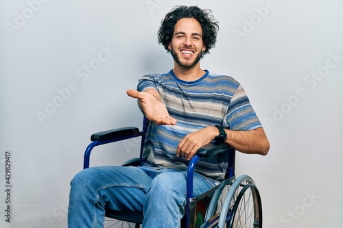 Handsome hispanic man sitting on wheelchair smiling cheerful offering palm hand giving assistance and acceptance.