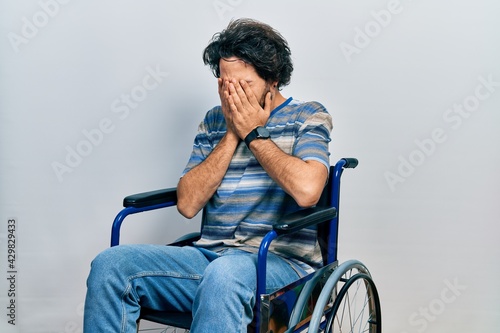 Handsome hispanic man sitting on wheelchair with sad expression covering face with hands while crying. depression concept.