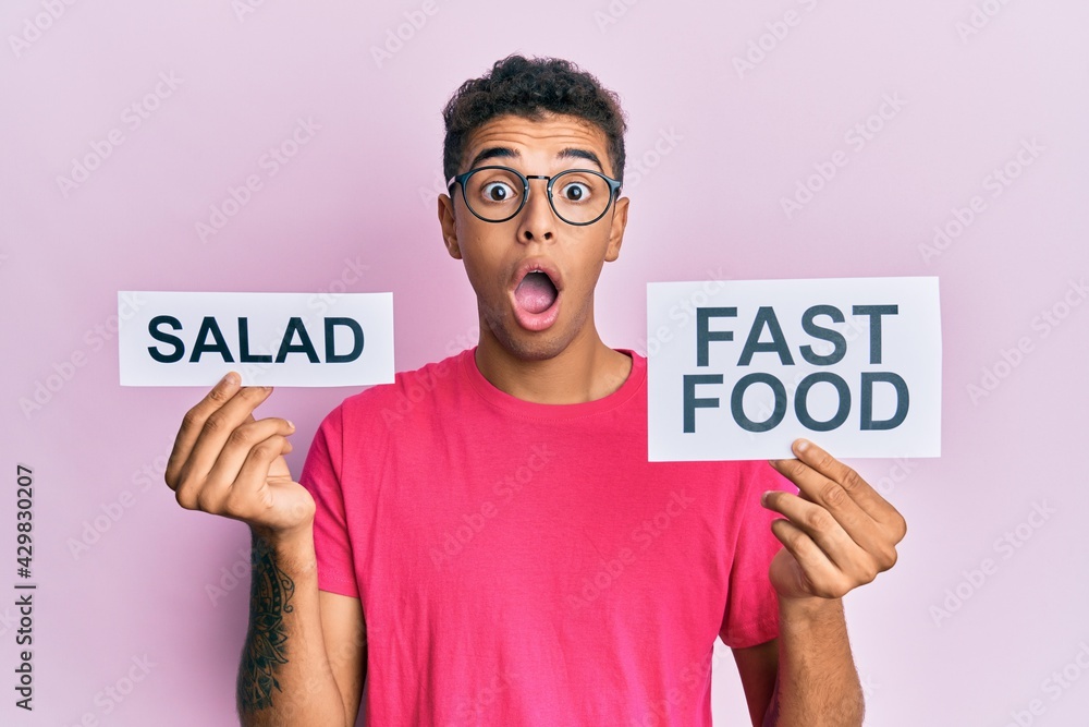 Young handsome african american man holding salad and fast food message paper afraid and shocked with surprise and amazed expression, fear and excited face. Young handsome african american man holding salad and fast food message paper afraid and shocked with surprise and amazed expression, fear and excited face.