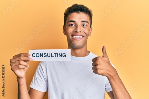 Young handsome african american man holding consulting message paper smiling happy and positive, thumb up doing excellent and approval sign © Krakenimages.com